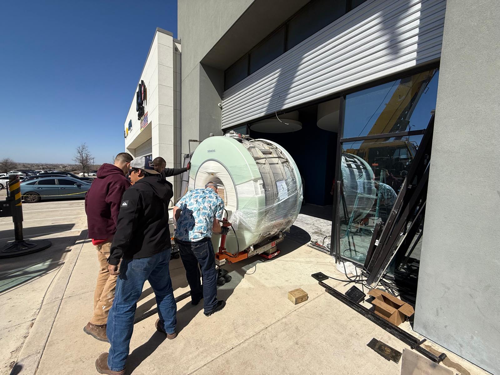Rigging crew guiding a shrink-wrapped Siemens MRI magnet on skates into a building through a removed storefront wall